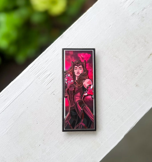 A rectangular enamel pin featuring an illustration of the Wanda in a dark red and black costume against a pink background, resting on a white painted wooden surface with blurred green foliage in the background.