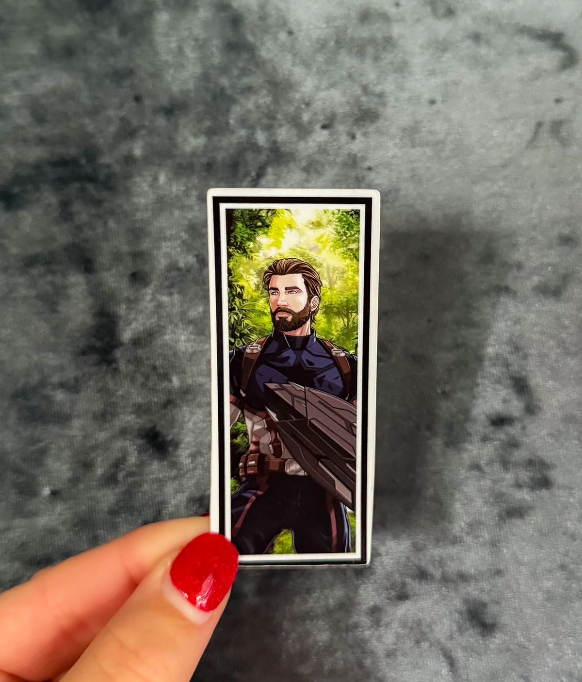 A rectangular printed card or sticker featuring an illustrated bearded man in a dark outfit against a green forest background, held between fingers with red nail polish against a mottled gray surface.