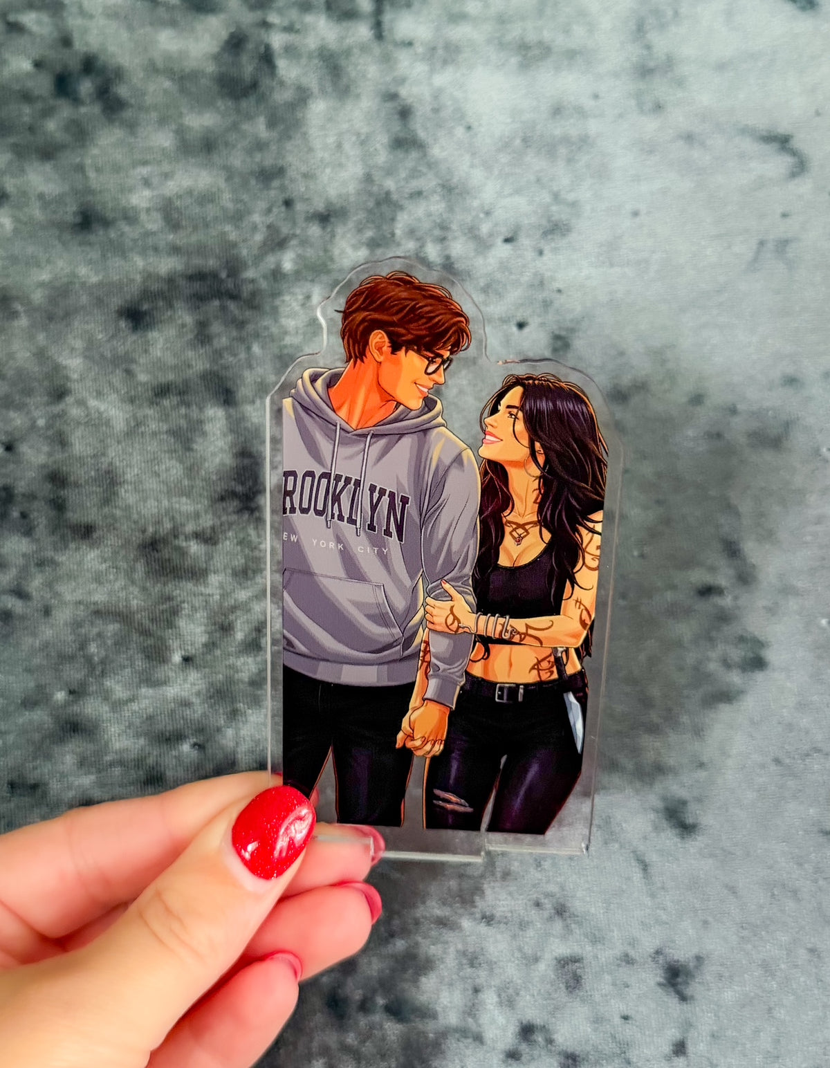 A clear acrylic standee featuring an illustrated couple, held by a hand with red nail polish against a textured gray background.