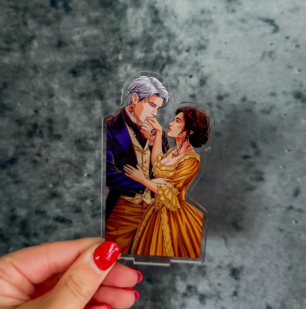 A hand with red nail polish holds a clear acrylic standee featuring an anime-style illustration of a silver-haired man in a dark blue suit and a woman in a gold dress, against a textured gray background.