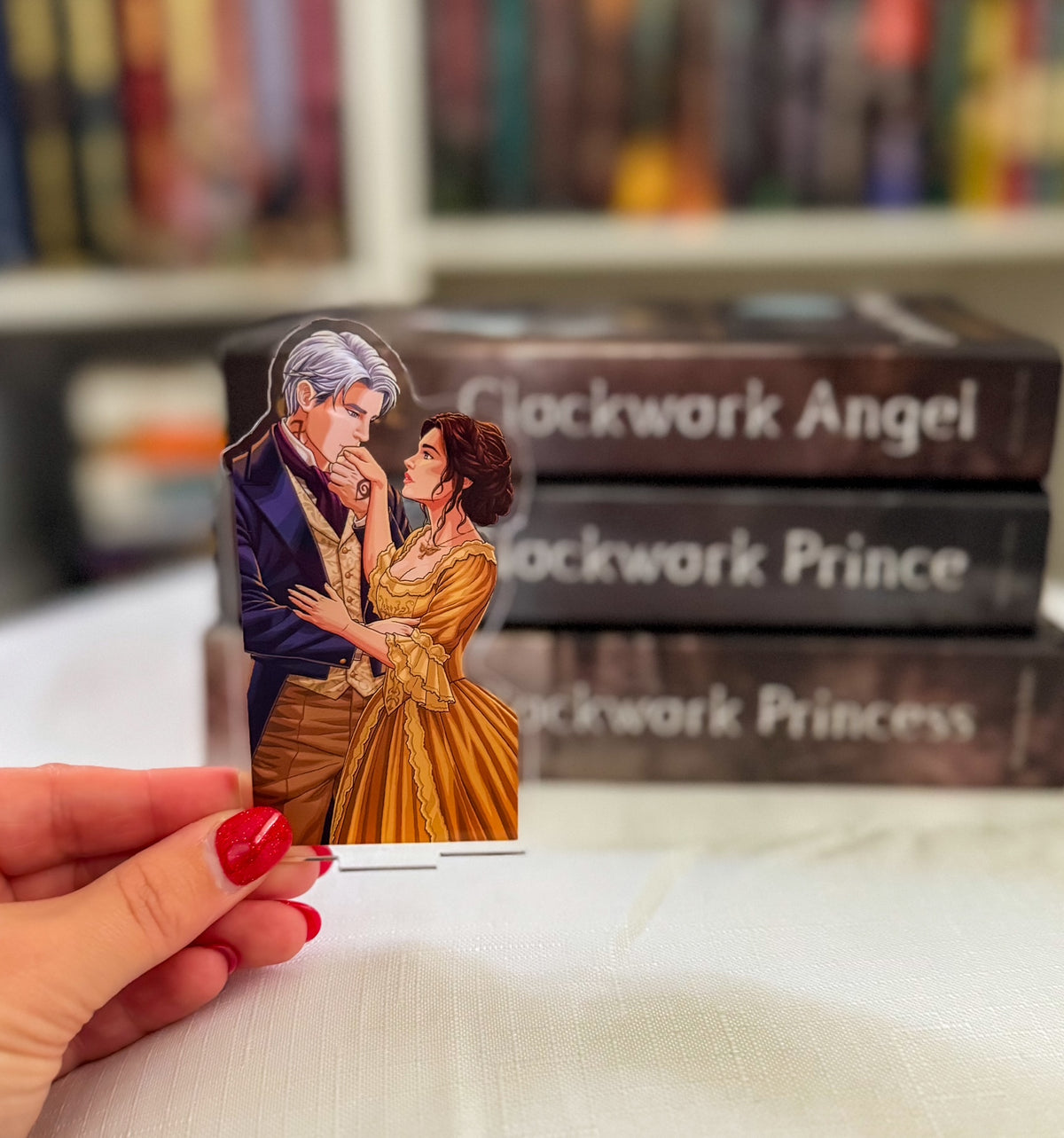 A hand with red nail polish holds a die-cut acrylic standee featuring an illustration of a man and a woman in period attire, with blurred book boxes in the background labeled 'Clockwork Angel', 'Clockwork Prince', and 'Clockwork Princess'.
