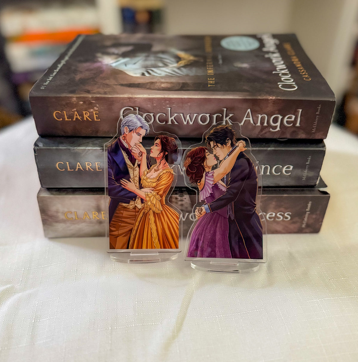 Two acrylic book-themed bookmarks featuring characters from 'Clockwork Angel' by Clare, displayed in front of a stack of the physical book covers on a white tablecloth.
