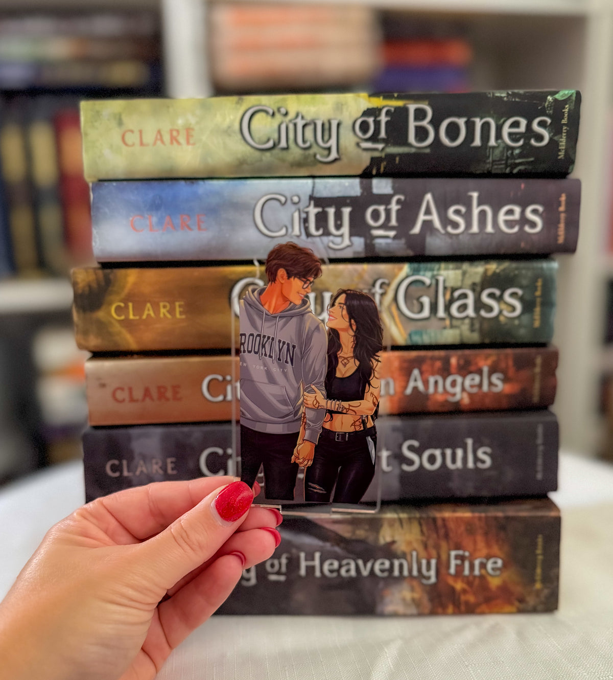 A hand with red nail polish holds a clear acrylic book charm featuring an illustration of a couple, positioned in front of a stack of seven novels by Clare. The books are arranged vertically on a white surface with a blurred bookshelf background.