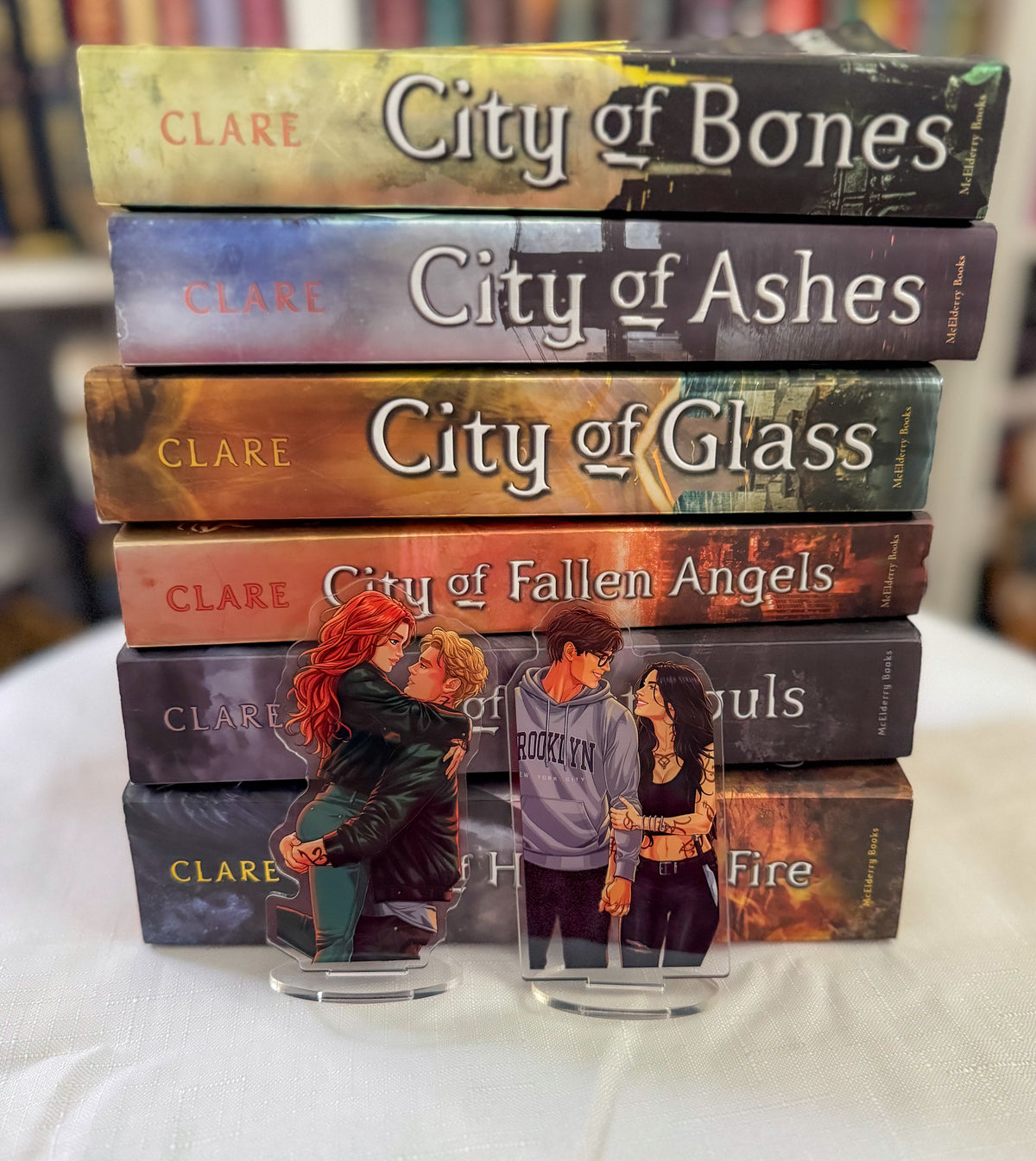 A stack of six hardcover books by Clare on a white tablecloth, with two acrylic character stands in front. The books are titled City of Bones, City of Ashes, City of Glass, City of Fallen Angels, and City of Fire.