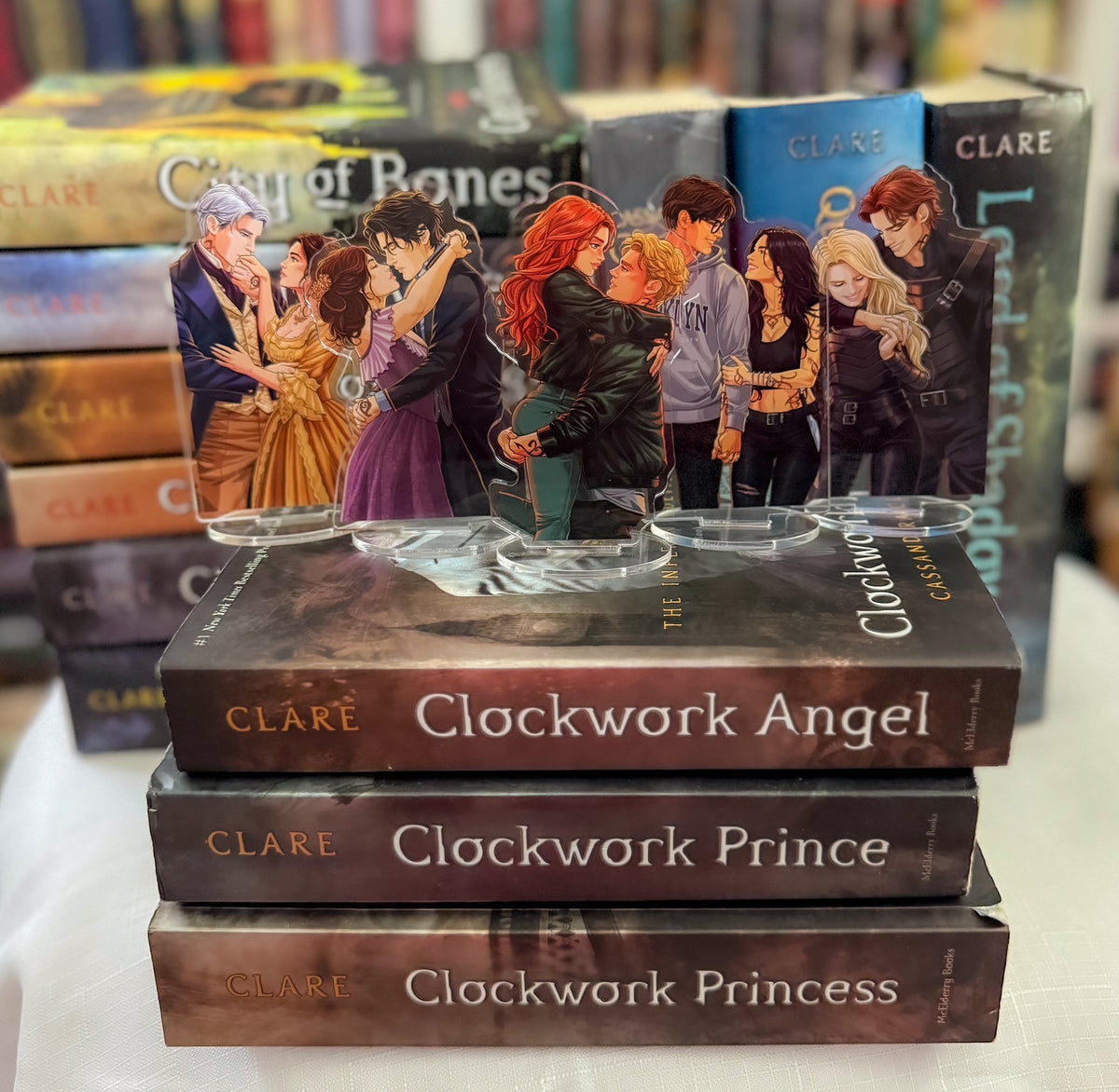 A set of clear acrylic book-themed stands featuring characters from the novels, displayed on top of stacked books including 'Clockwork Angel', 'Clockwork Prince', and 'Clockwork Princess' by Clare.