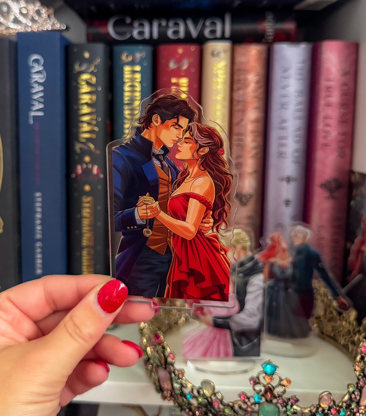 A hand with red nail polish holds a die-cut acrylic standee featuring an illustration of a romantic couple embracing, set against a background of books and jewelry.