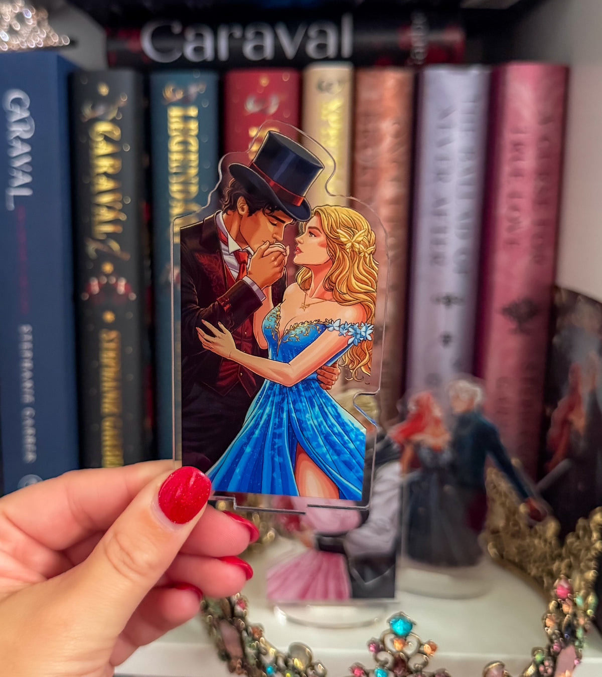 A hand with red nail polish holds a clear acrylic standee featuring an illustrated couple embracing, with a blonde woman in a bright blue dress and a dark-haired man in a top hat. The background shows a bookshelf with books and other collectible figures.