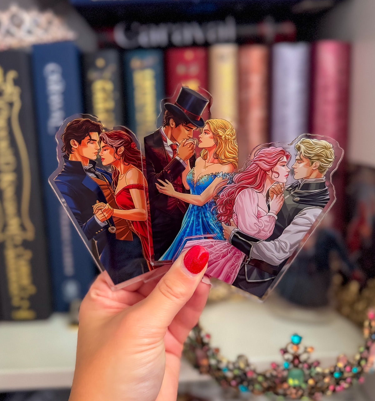 A hand with red nail polish holds a set of four die-cut acrylic bookmarks or tags featuring illustrated romantic couple scenes, with blurred book spines in the background.