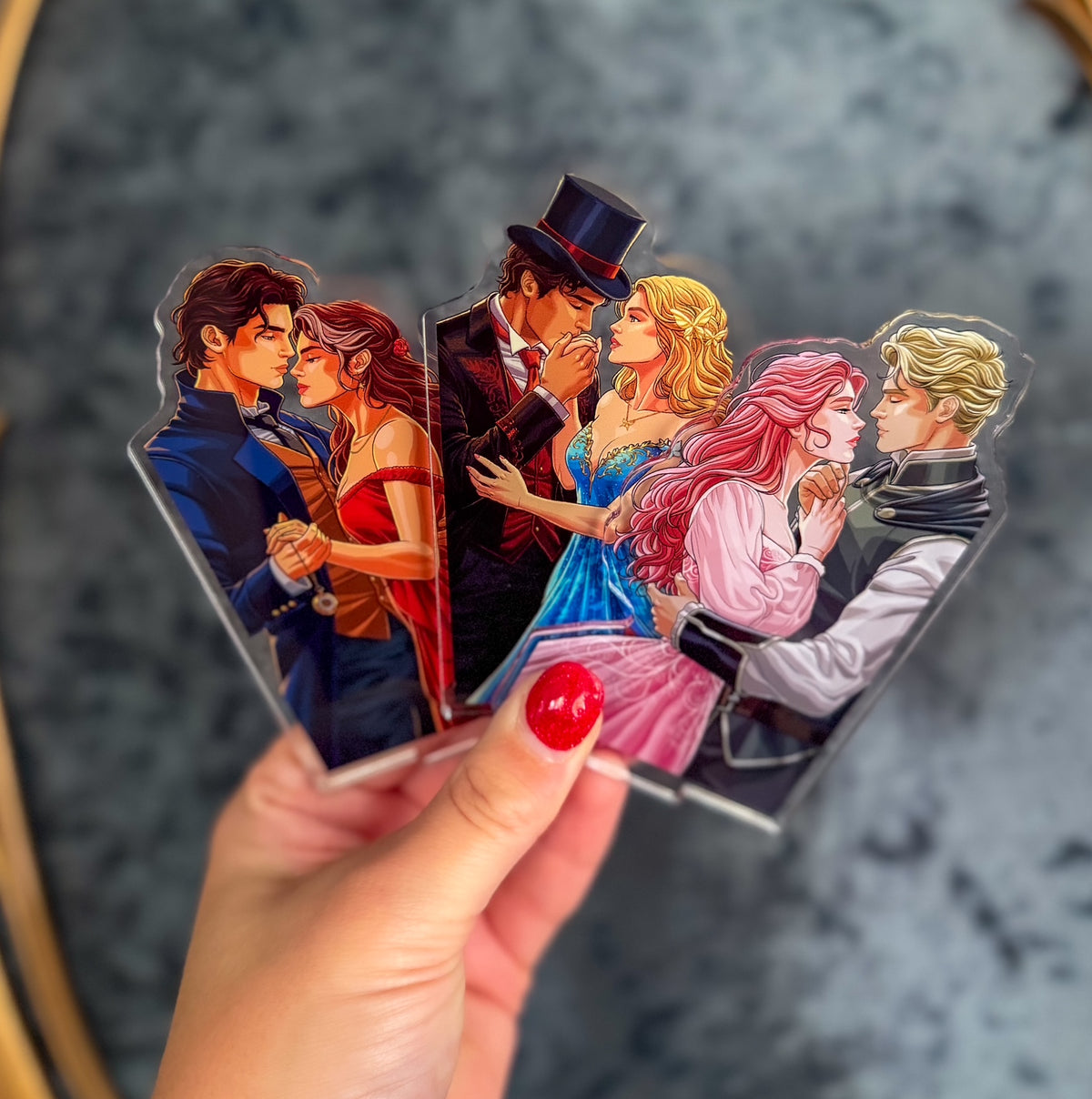 A hand with red painted fingernails holds three colorful acrylic standees featuring illustrated romantic couple scenes. The background is a blurred gray textured surface.