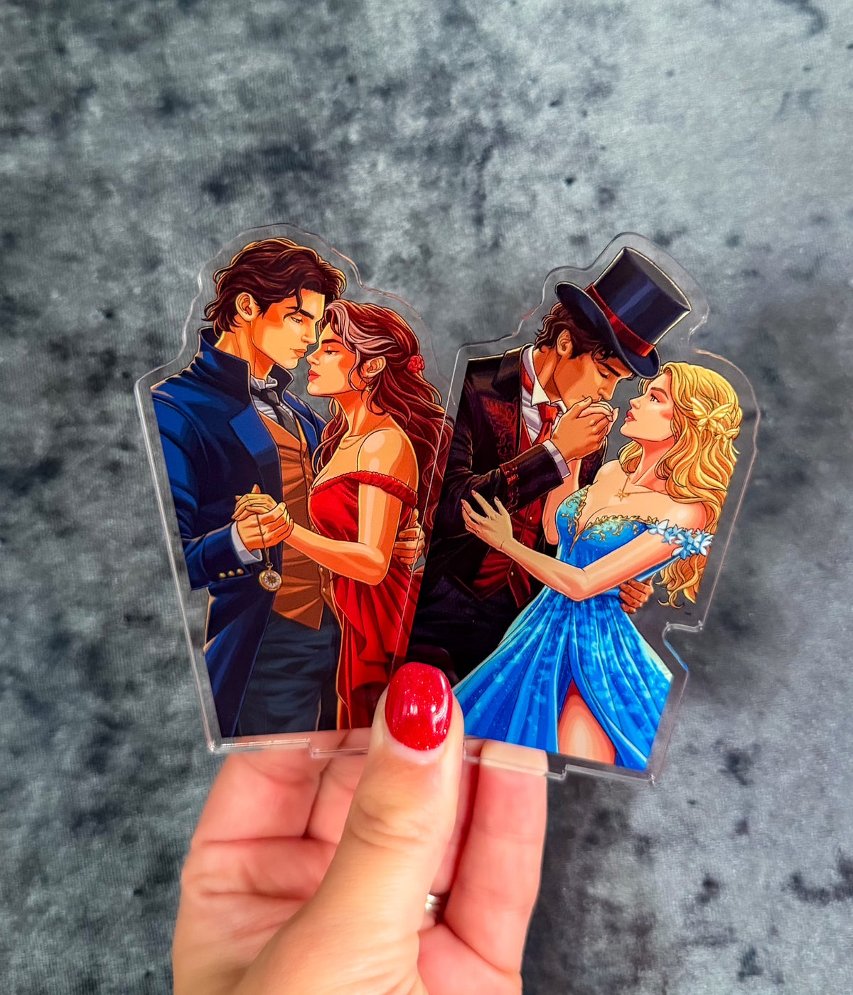 A hand with red nail polish holds two clear acrylic standees featuring illustrated couples. One couple wears blue and red formalwear, and the other wears black and blue formalwear.