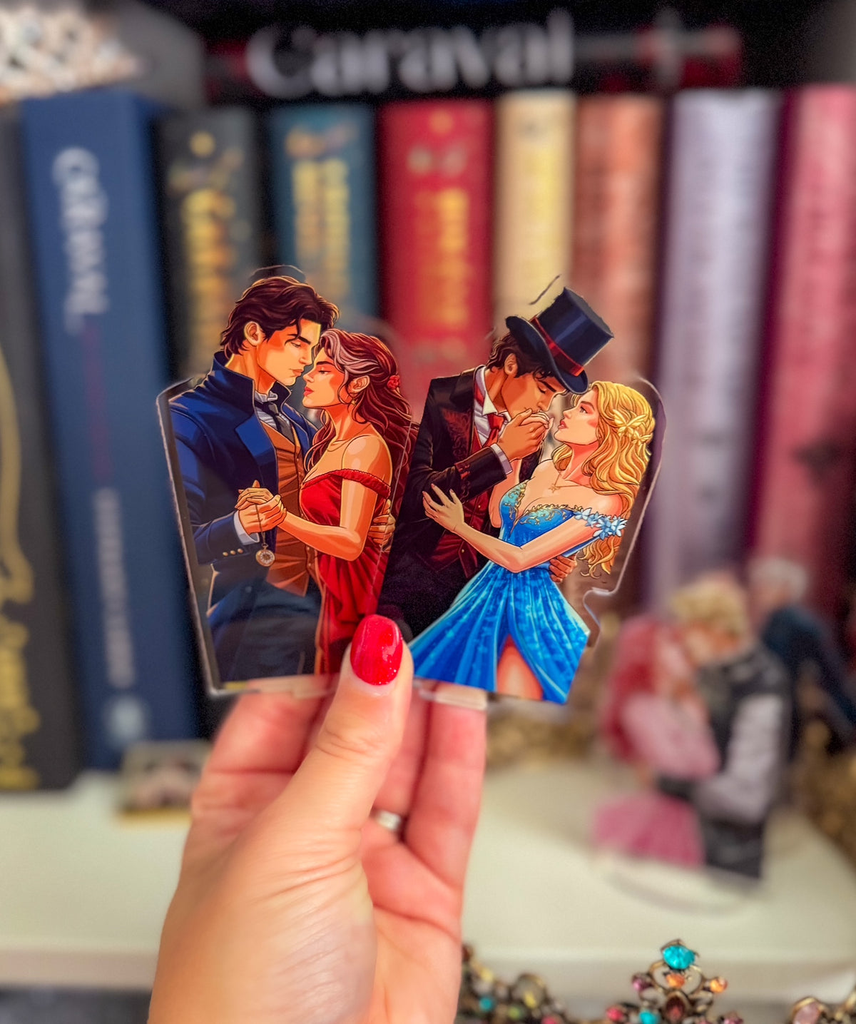 A hand with red nail polish holds a die-cut acrylic charm featuring two romantic couple illustrations, set against a blurred background of books and collectible figurines.