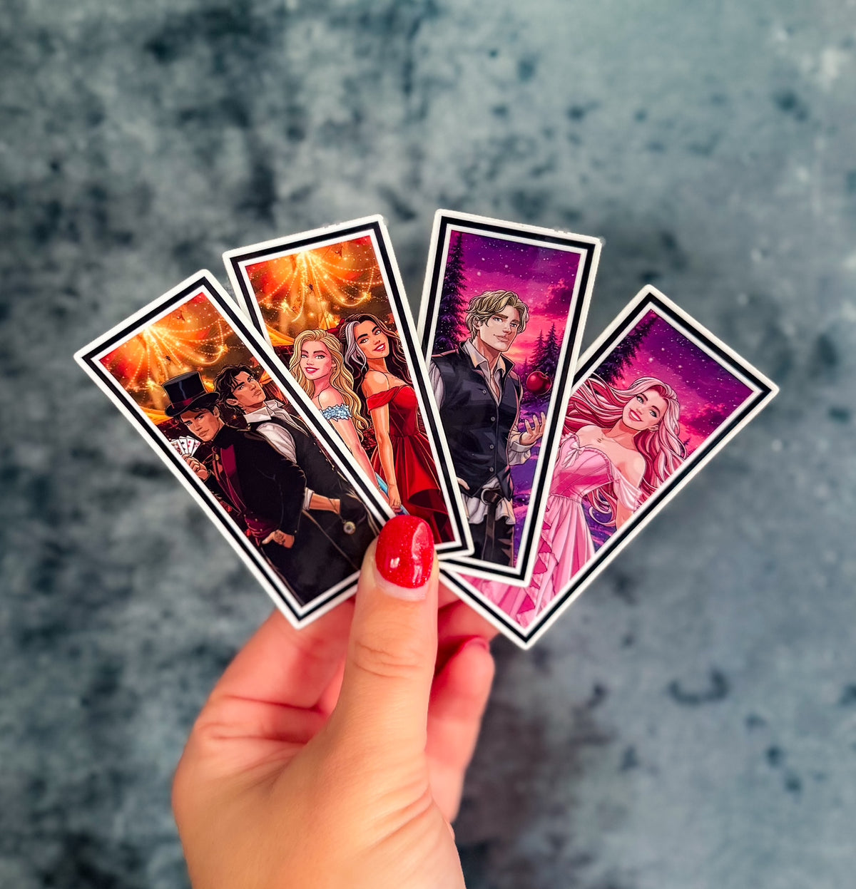 A hand with red nail polish holds four illustrated rectangular cards or stickers fanned out against a textured gray-blue background. The cards feature fantasy-style character artwork in vivid colors: fiery orange and black, red and black, pink and black, and purple and pink scenes.