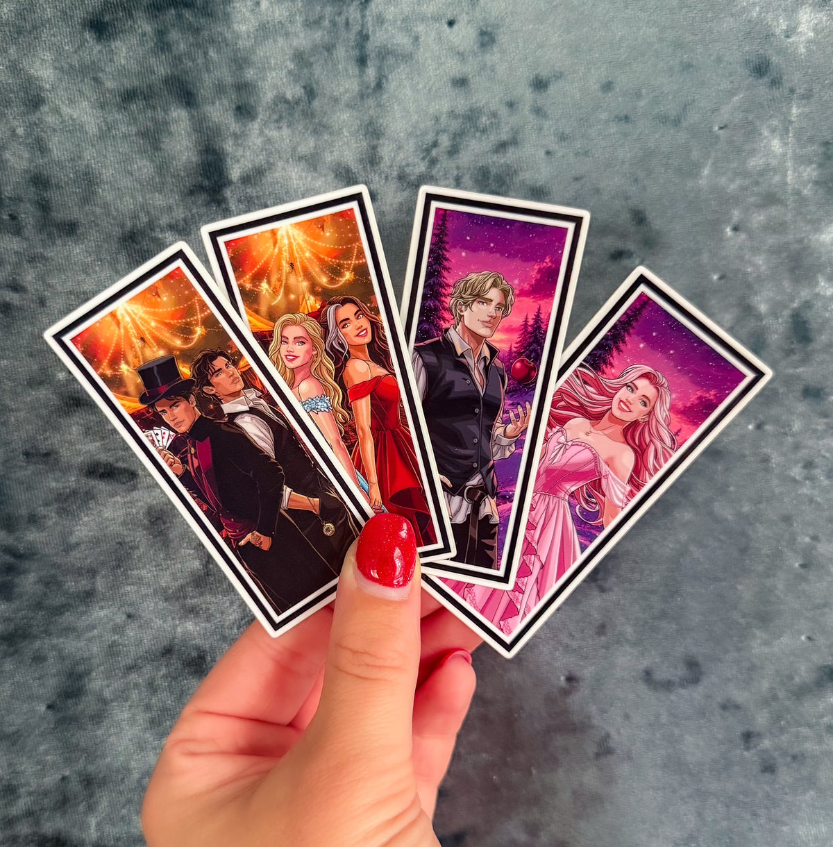 A hand with red painted fingernails holds four illustrated rectangular cards fanned out against a mottled gray background. The cards feature anime-style characters in colorful fantasy and dramatic scenes.