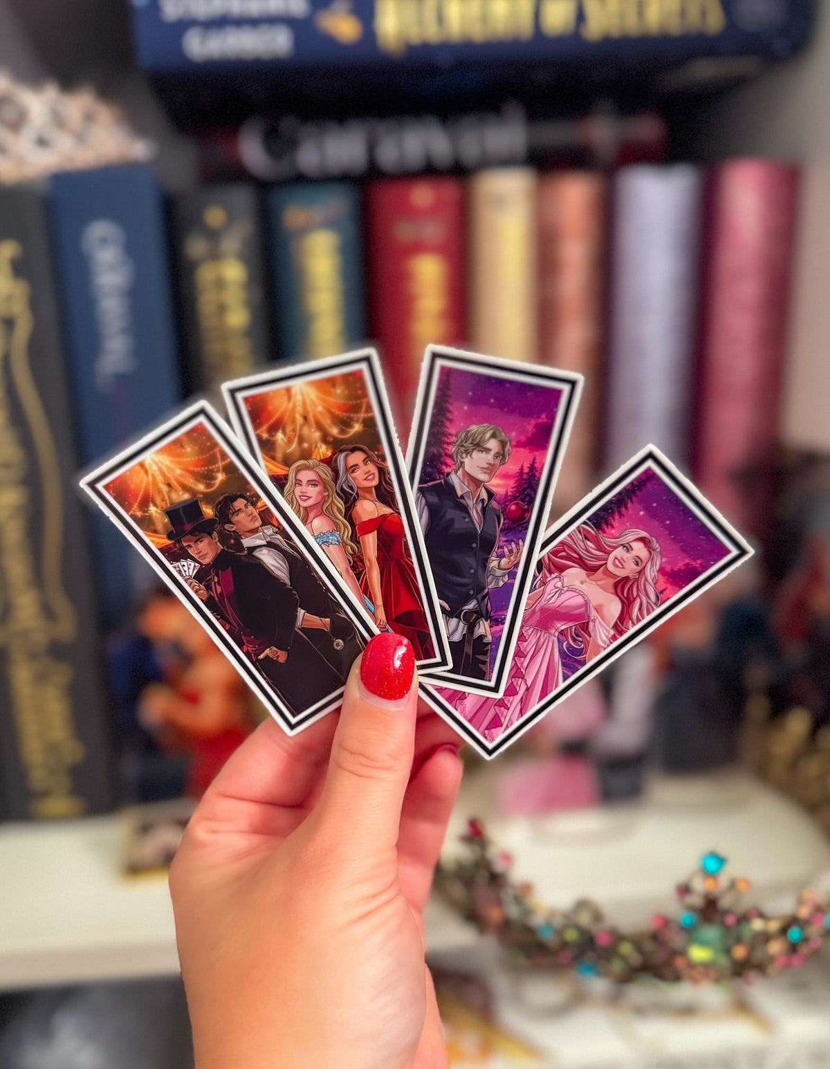 A hand with red nail polish holds four illustrated anime-style cards or stickers fanned out in front of a blurred background of books and decorative items.