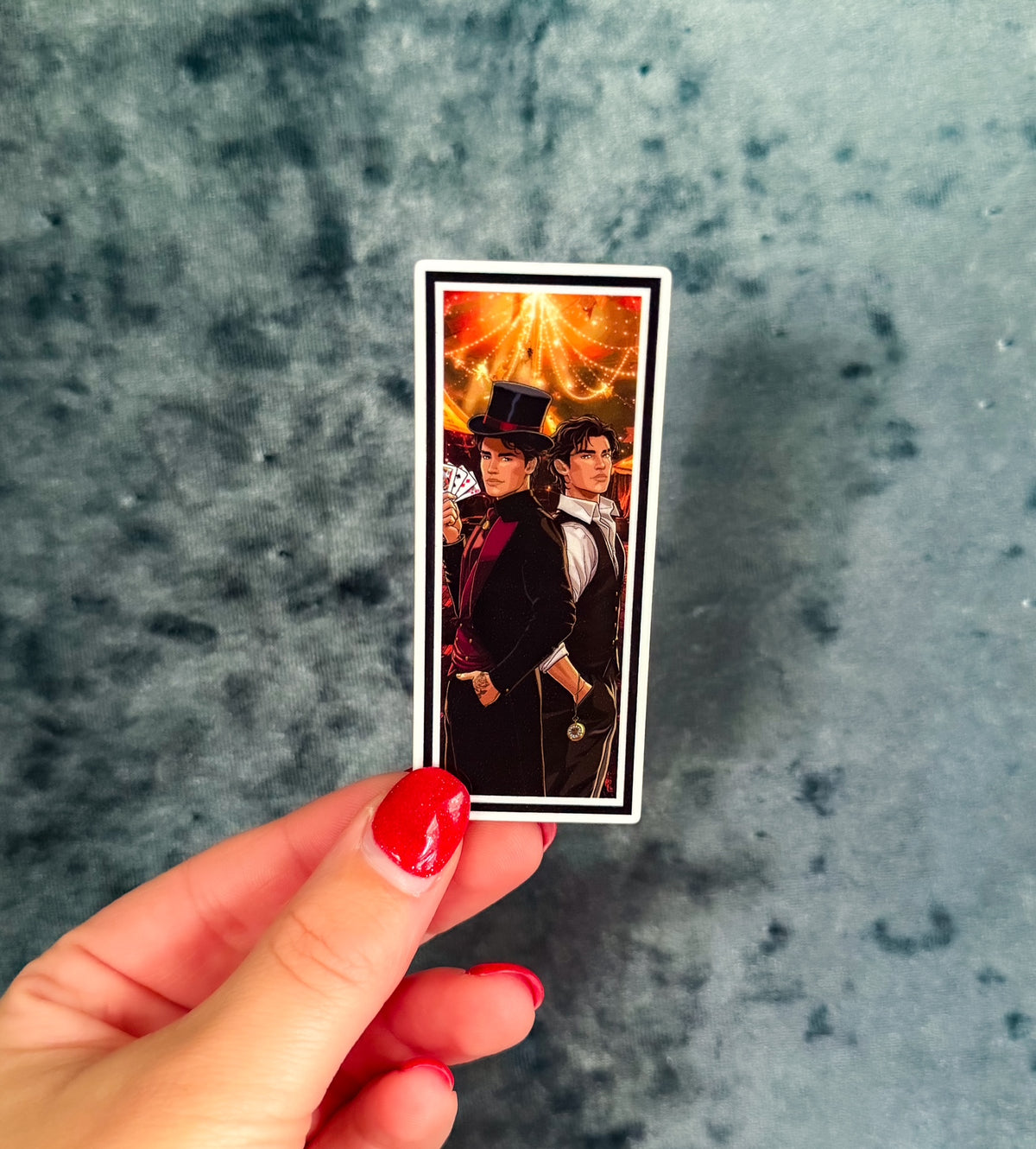 A hand with red nail polish holds a small rectangular printed card or sticker featuring a portrait of two men in formal black clothing against a warm glowing background, set against a mottled gray-blue surface.