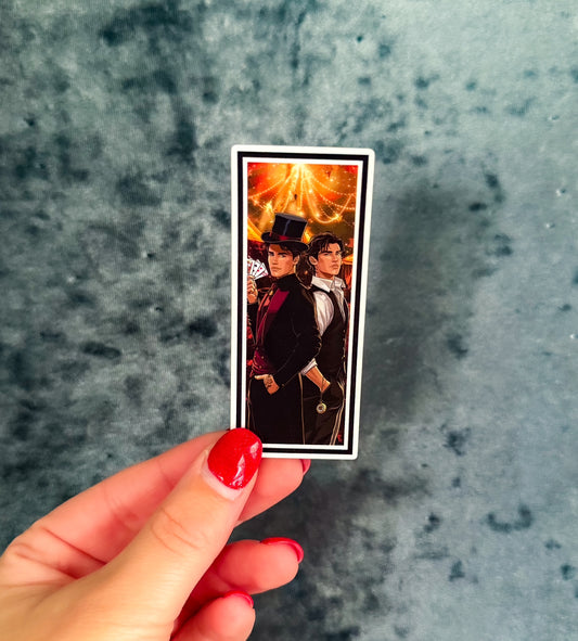 A hand with red nail polish holds a small rectangular printed card or sticker featuring a portrait of two men in formal black clothing against a warm glowing background, set against a mottled gray-blue surface.