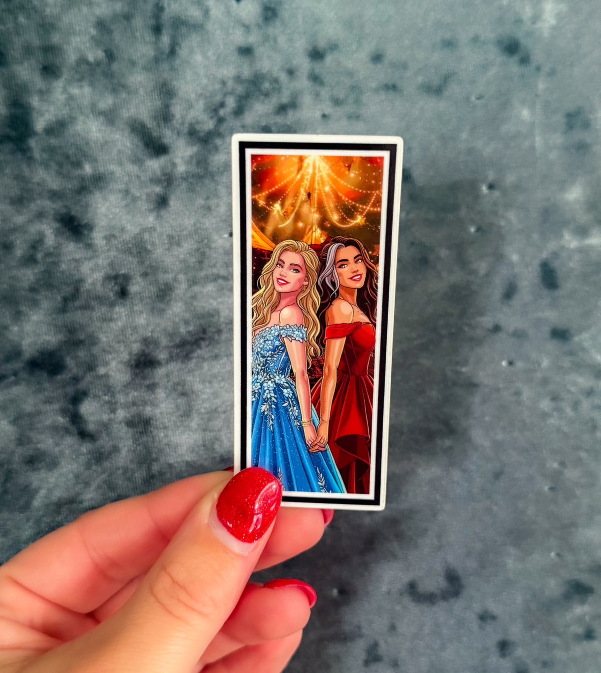 A hand with red nail polish holds a rectangular printed card or sticker featuring an illustrated scene of two women in formal dresses under a glowing orange sky, set against a mottled gray-blue background.
