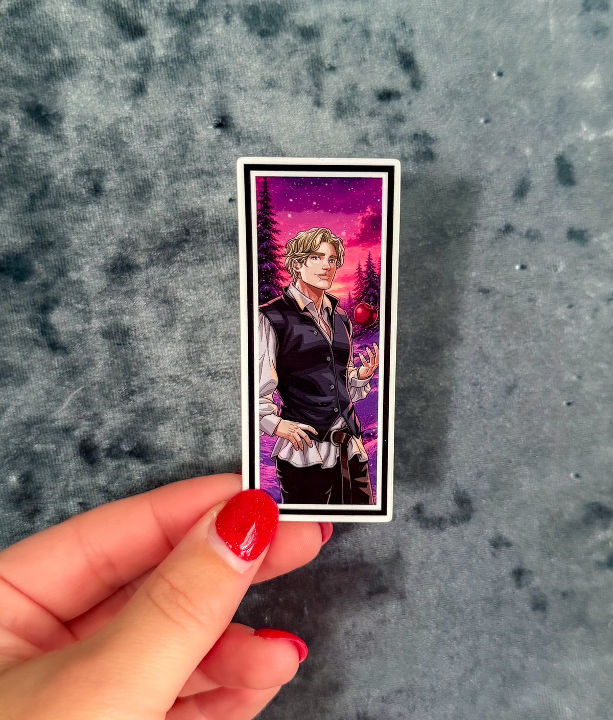 A hand with red nail polish holds a rectangular printed card or sticker featuring an anime-style illustration of a blond man in a dark vest against a pink and purple night sky, set against a mottled gray background.