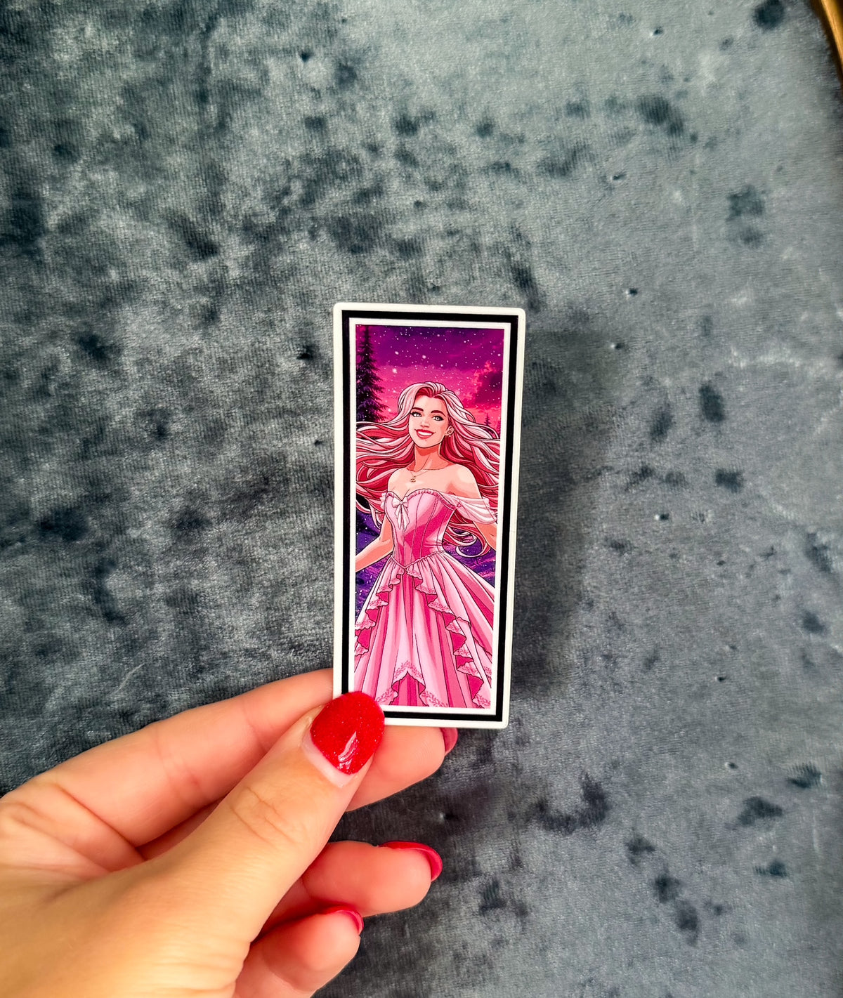 A hand with red nail polish holds a small rectangular printed card or sticker featuring an illustrated woman in a pink gown against a purple and pink starry sky. The background is a textured gray surface.