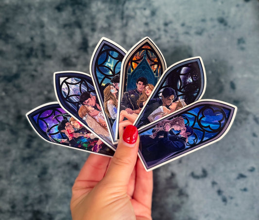 A hand with red nail polish holds a fan of five die-cut stickers featuring illustrated gothic window scenes in blue, purple, and black tones, set against a textured gray background.