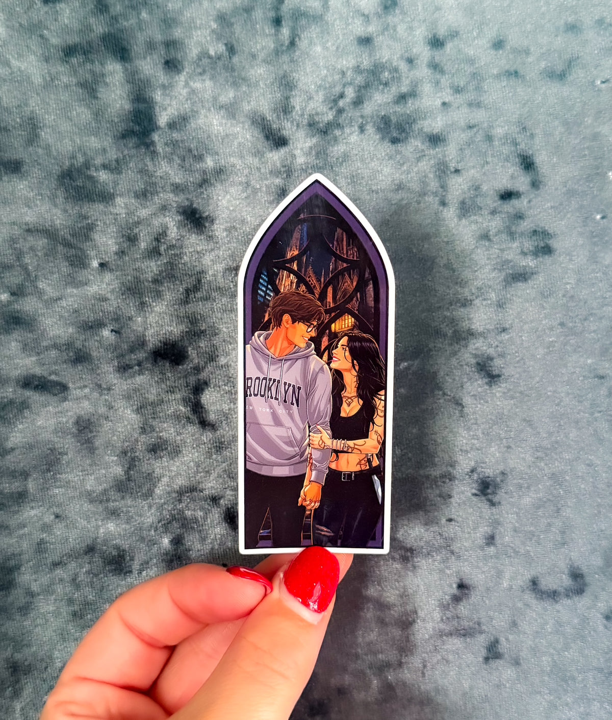 A hand with red nail polish holds a die-cut sticker shaped like a gothic arch window, showing an illustrated couple embracing. The sticker is placed on a textured gray surface.