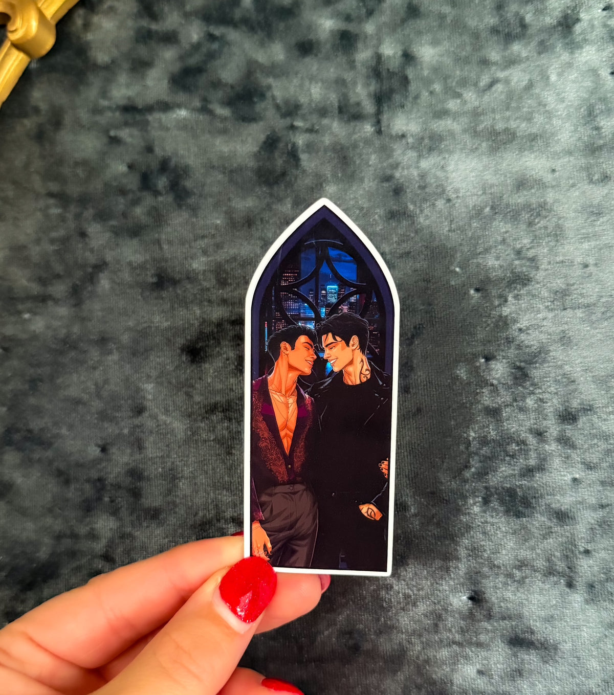 A hand with red nail polish holds a small arched sticker or magnet featuring an illustrated couple under a stained-glass-like window, set against a dark textured gray background.