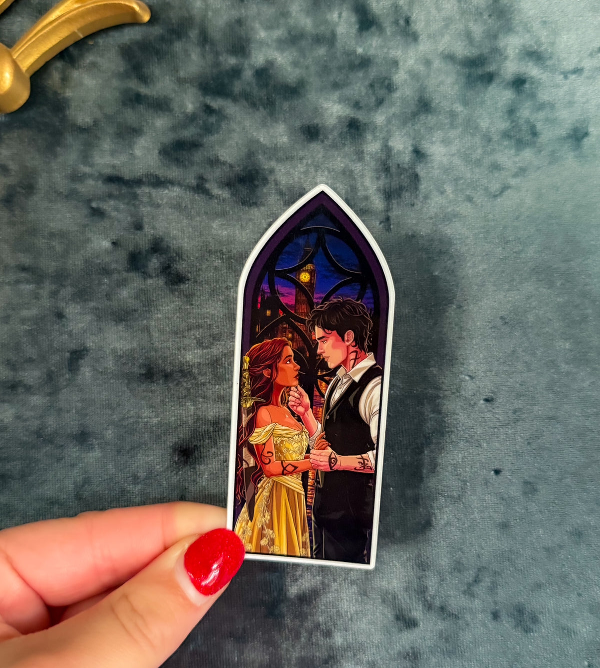 A hand with red nail polish holds a die-cut arched sticker or magnet featuring an illustrated couple in a stained-glass window design, set against a dark textured gray background.