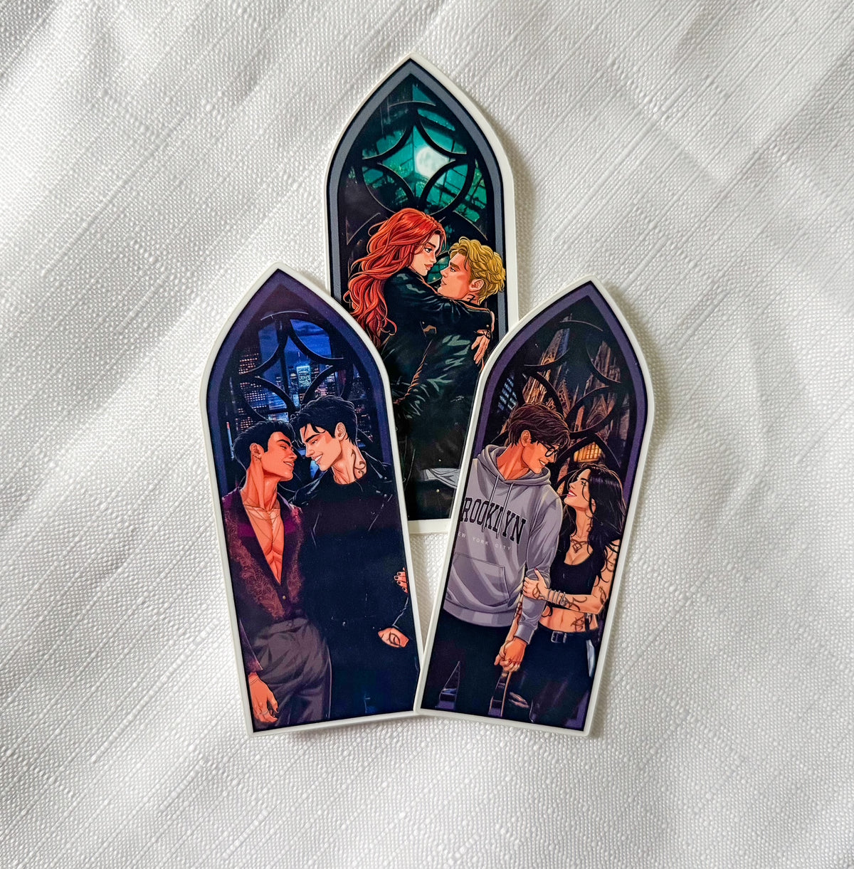 Three arched window-shaped stickers featuring illustrated couples in gothic stained-glass scenes, displayed on a white textured fabric background.