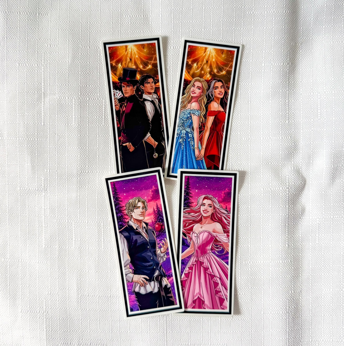 Four illustrated anime-style bookmarks or card sleeves arranged on a white textured fabric background. Each piece features a different character with colorful fantasy or formal-clothing artwork, including orange/gold, blue/red, and pink/purple scenes.